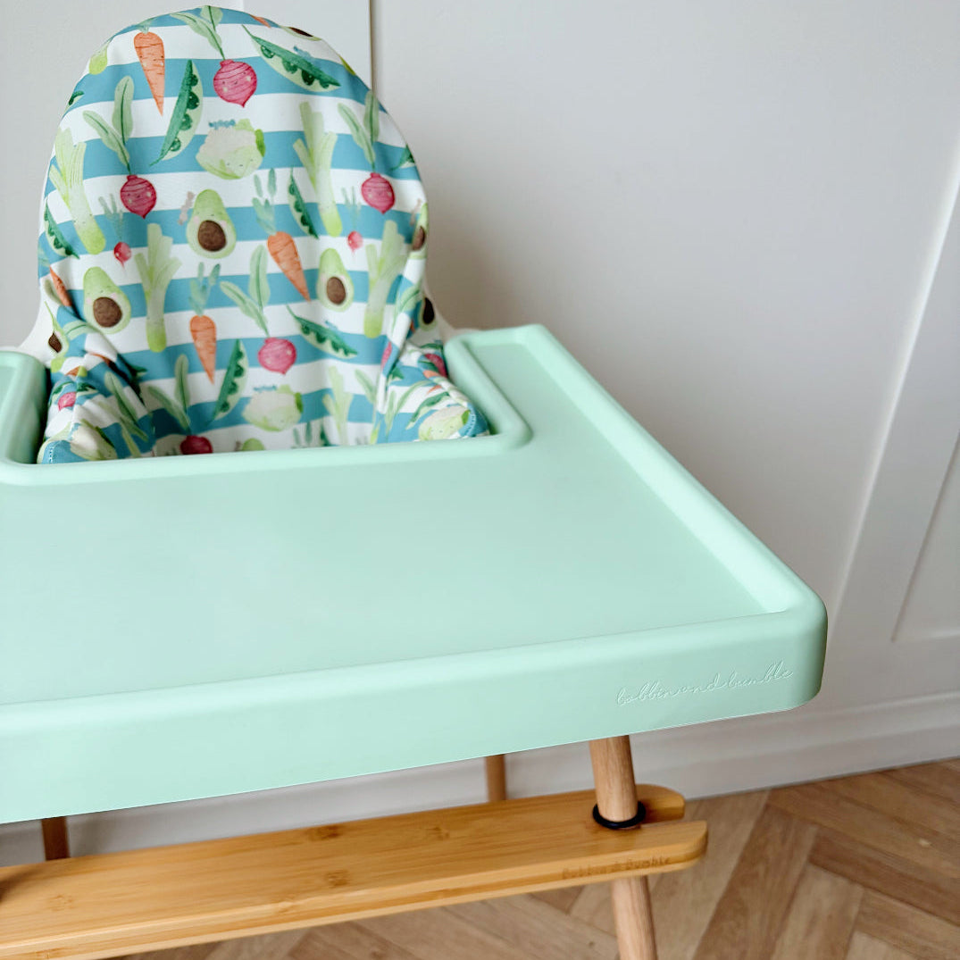 Mint green high chair tray cover with a colourful cushion on a wooden floor.