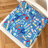 A square blue chair cushion with a colorful craft supply pattern, shown on a white chair over a wooden parquet floor.