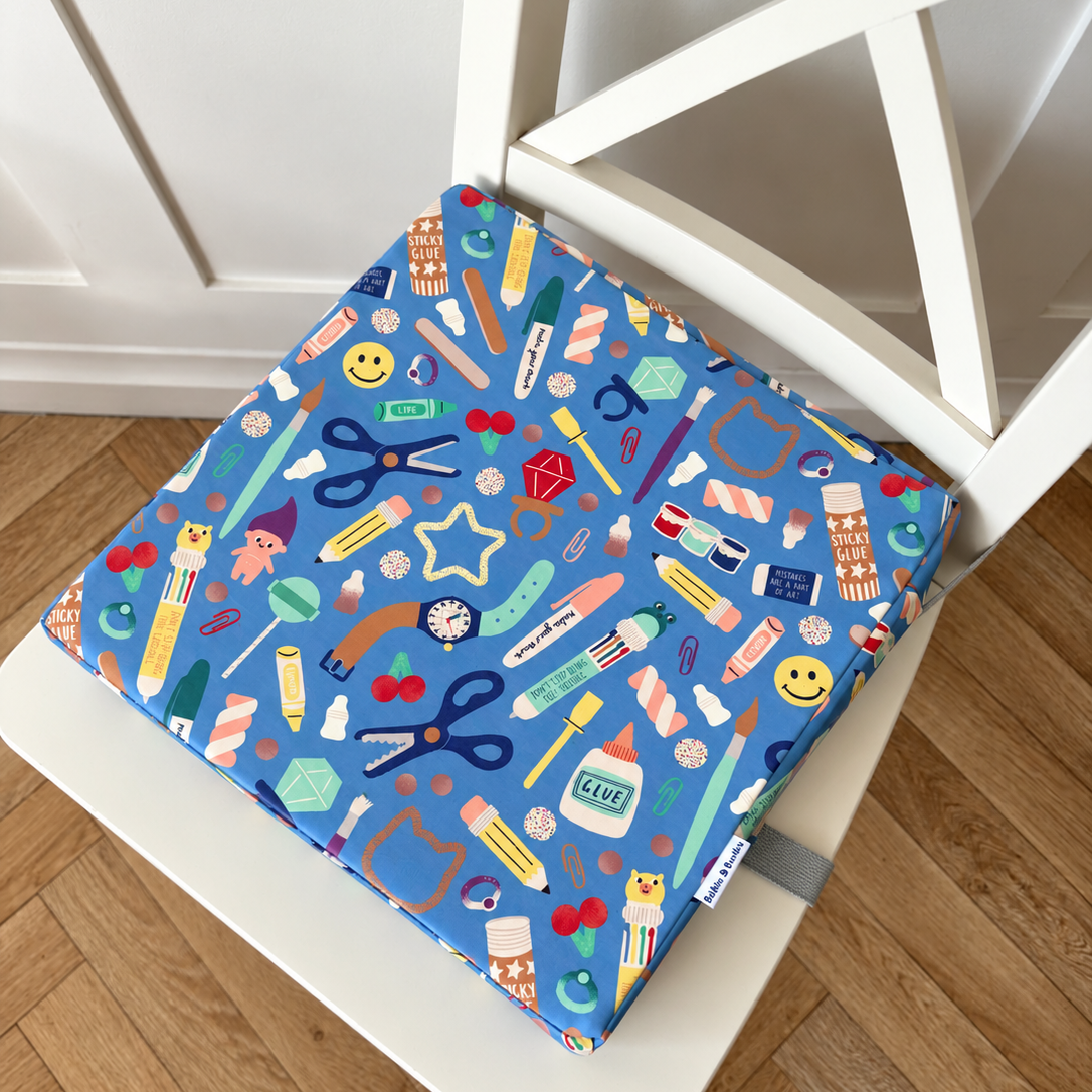 A square fabric chair cushion with a colourful craft supply pattern, secured on a white wooden chair over a light brown wood floor.