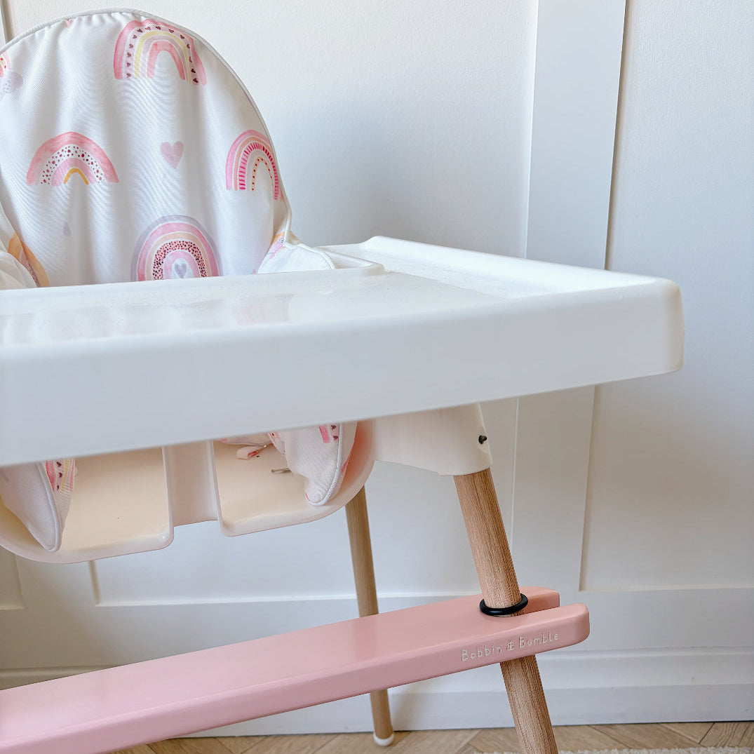 White high chair with pink footrest and rainbow-patterned cover on a wooden floor.
