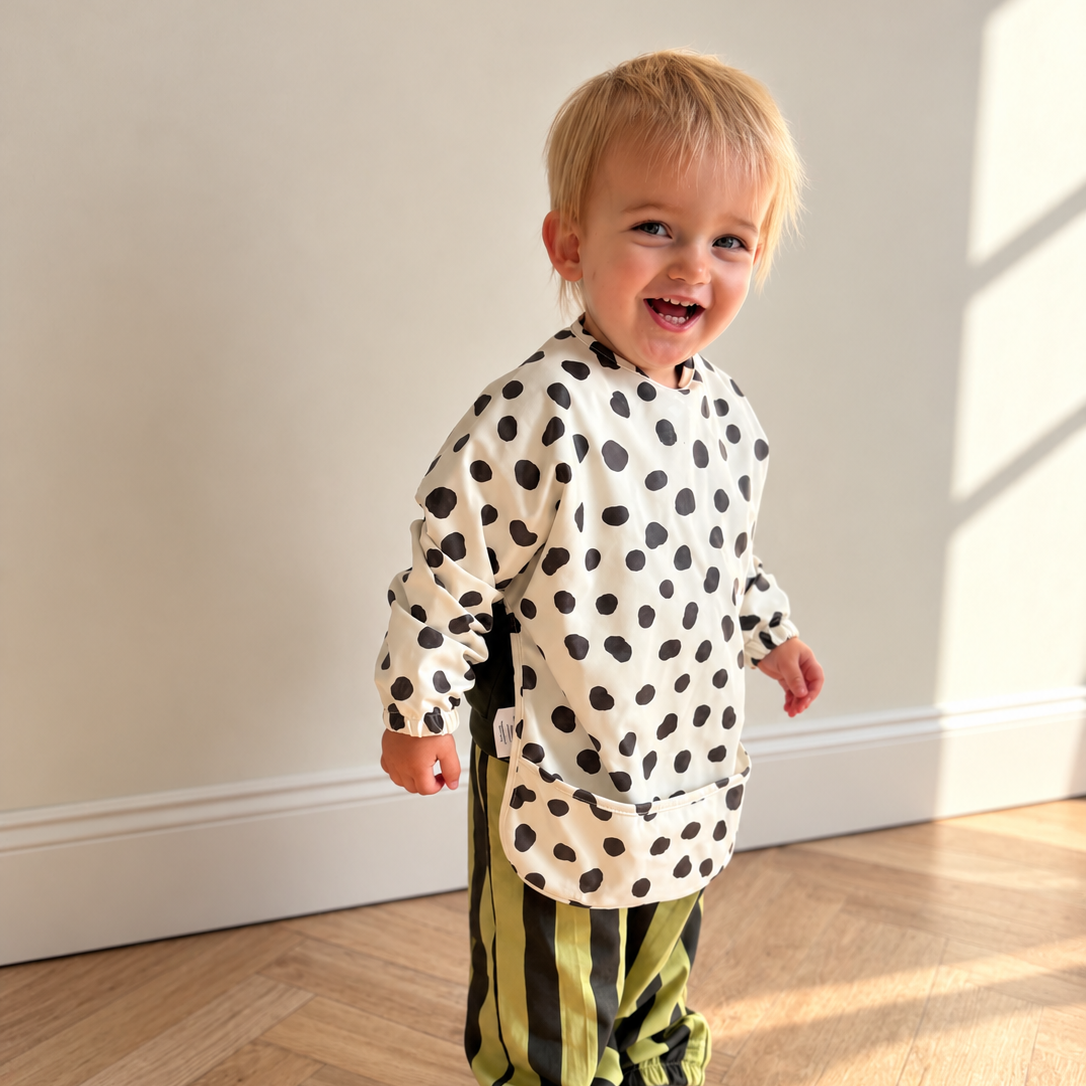 A toddler standing in a sunlit room with a light wood floor and cream wall, wearing a cream long-sleeve bib with black spots and olive green striped pants.