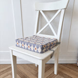 A square fabric booster cushion with a blue, white, and mustard geometric diamond pattern, shown resting on a white wooden chair in a room with a light wood herringbone floor.