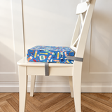 A blue booster cushion with a colourful crafting pattern and grey straps, placed on a white wooden chair in front of a white paneled wall and wood herringbone floor.