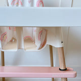 Children's high chair with rainbow cushion, pink footrest and wooden legs on a light wood floor.