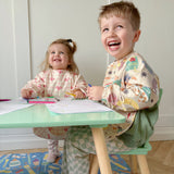 Two children sitting at a table with colourful long-sleeved bibs and crayons, smiling.