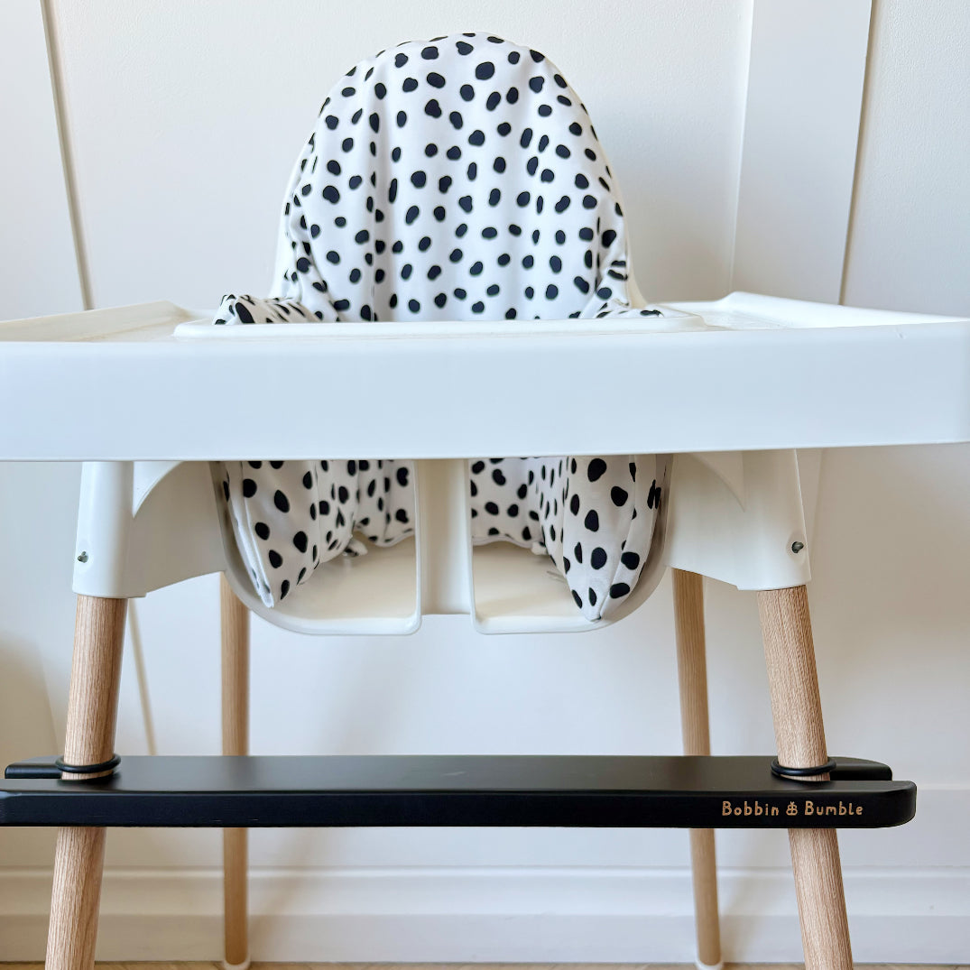 White high chair with black spotty seat cushion and wooden legs with a black footrest on a light wood floor.
