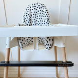 White high chair with black spotty seat cushion and wooden legs with a black footrest on a light wood floor.
