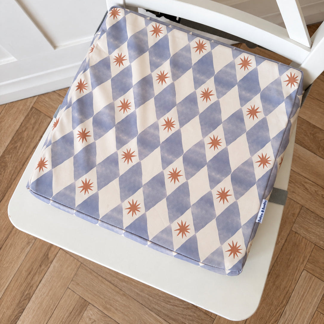 A square toddler booster cushion with a blue and cream harlequin pattern and small yellow starburst accents, shown resting on a white chair over a wooden floor.