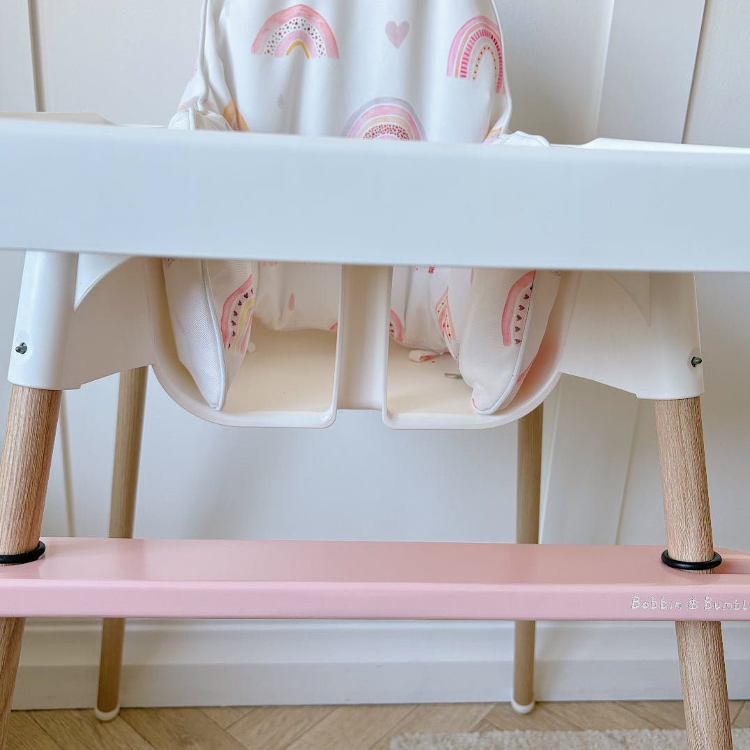 White high chair with pink seat and rainbow-patterned cover against a white wall.