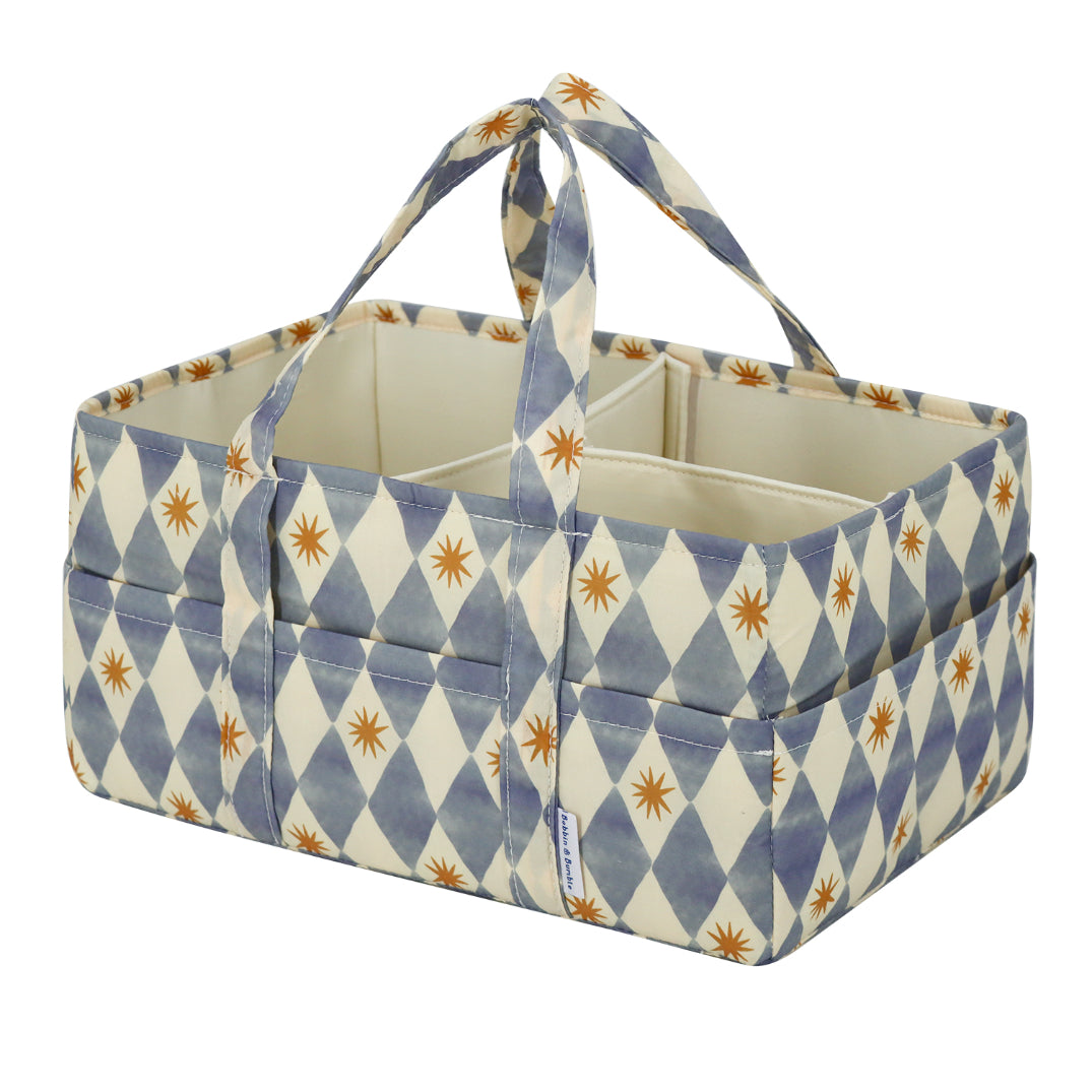 A fabric nappy caddy basket with a blue and cream harlequin diamond pattern accented by small mustard starburst motifs. The basket features multiple front pockets and two top handles, shown on a solid white background.
