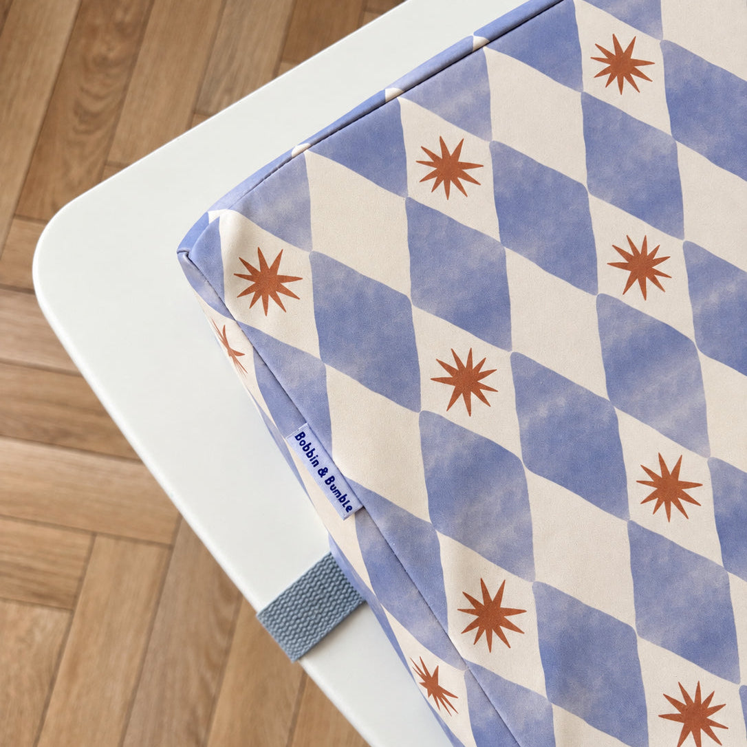 A close-up of a toddler booster cushion with a blue-and-white harlequin pattern and yellow starburst motifs, resting on a white table over a wooden floor. A small fabric Bobbin and Bumble Tag