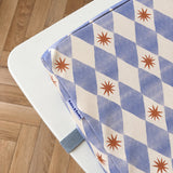 A close-up of a toddler booster cushion with a blue-and-white harlequin pattern and yellow starburst motifs, resting on a white table over a wooden floor. A small fabric Bobbin and Bumble Tag