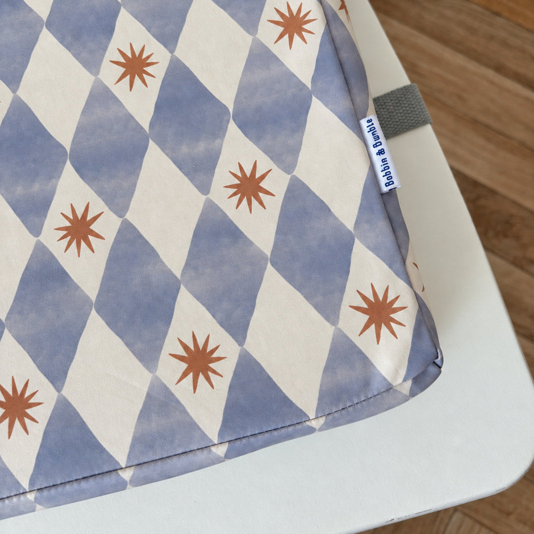 A close-up of the Uppy toddler booster cushion with a blue and cream harlequin diamond pattern and brown starburst motifs, resting on a white table with a wooden floor in the background.