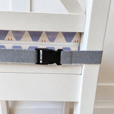 A gray webbed safety restraint strap with a black plastic buckle, shown attached to a white wooden chair in a home setting with a wood floor visible.
