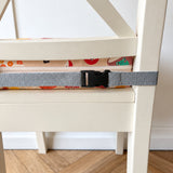 A close-up of a gray adjustable seat belt with a black buckle strapped across the seat of a cream-colored wooden chair. The chair is set against a white wall and light wood herringbone floor.