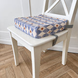 A square toddler booster cushion with a blue, cream, and yellow diamond pattern sits on a white wooden chair in a room with a light wood herringbone floor.