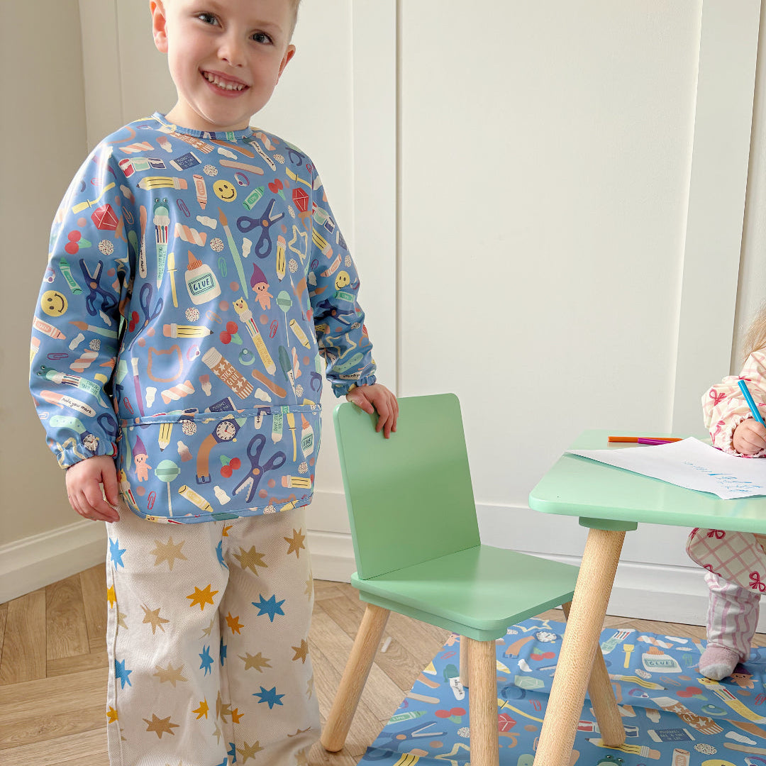 Child wearing a colorful outfit with crafting items design, standing next to a small table and chair in a room.