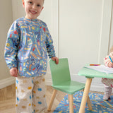 Child wearing a colorful outfit with crafting items design, standing next to a small table and chair in a room.