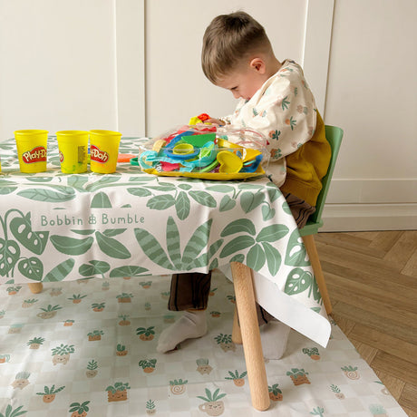 Child sat at a table playing with play-doh. Plant pot splash mat underneath and a leafy green splash mat used as a tablecloth