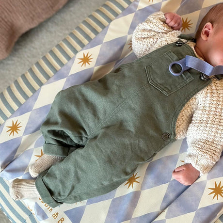 Baby lying on a harlequin patterned changing mat wearing green overalls and a knit sweater.