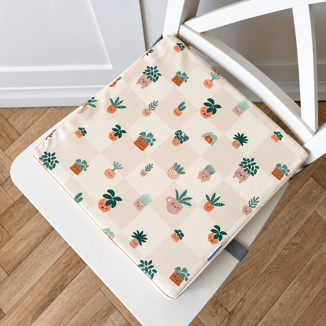 A square booster seat cushion with a cute potted plant pattern, displayed on a white wooden chair over a light wood herringbone floor.