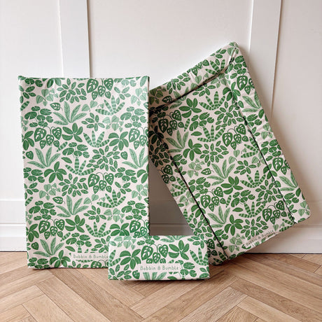 Three baby changing mats with a green botanical leaf pattern on a cream background, arranged on a light wood herringbone floor against a white panelled wall.