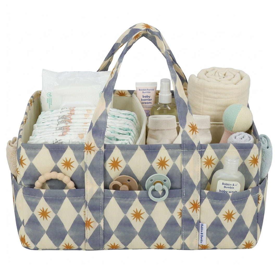 A fabric baby nappy caddy organizer with a blue and cream harlequin pattern and gold starburst motifs, shown filled with baby essentials including nappies, bottles, a rolled towel, a dummy, and teething toys on a white background.