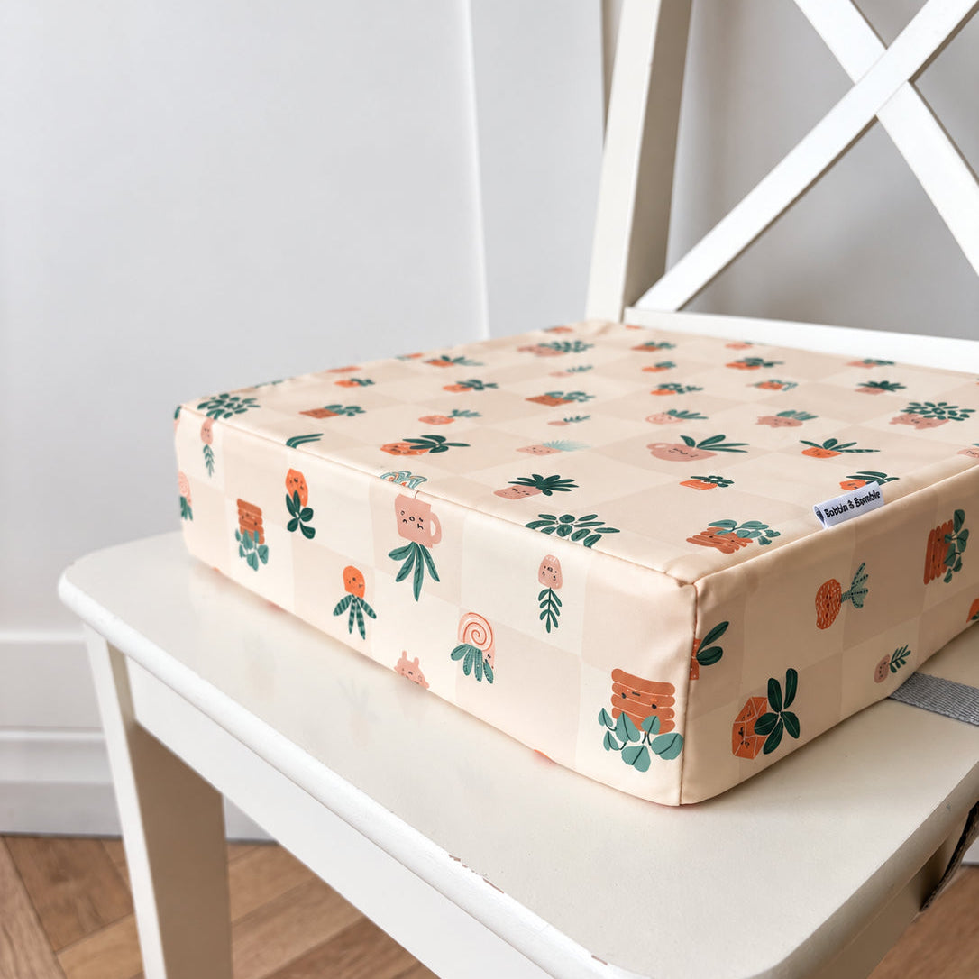 A square booster cushion with a light beige background and a colourful pattern of cartoon plants, resting on a white wooden chair.