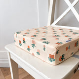 A square booster cushion with a light beige background and a colourful pattern of cartoon plants, resting on a white wooden chair.