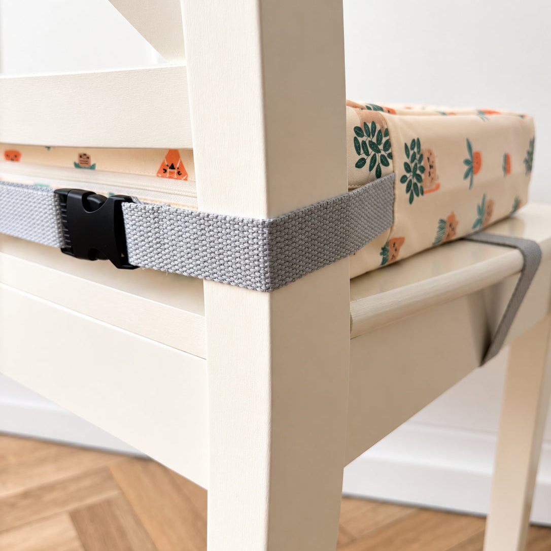 A close-up of a grey adjustable seat strap with a black plastic buckle, securing a patterned booster cushion on a white wooden chair in a home setting.