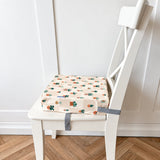 A square fabric chair booster cushion with a colourful plant pattern on a cream background, secured with a grey strap to a white wooden chair on a herringbone wood floor.