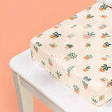 A square child's booster cushion with a cute potted plant pattern, resting on a white chair against a peach-colored background.