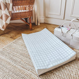 Pale green gingham changing mat in living room with various baby items including a wooden crib on a wooden floor 