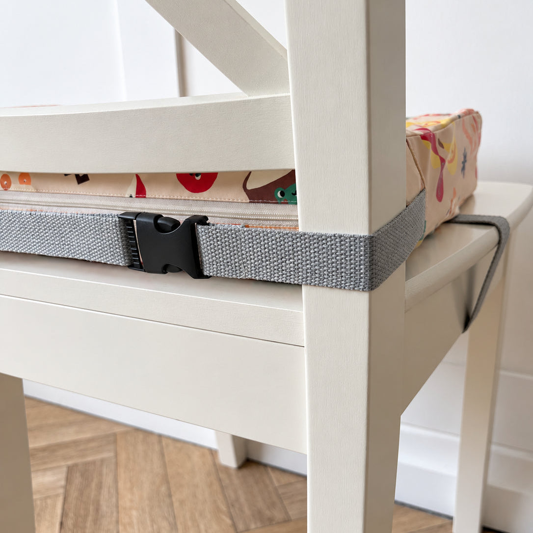 A grey woven seat belt with a black plastic buckle strapped across the seat of a white wooden chair, securing a patterned booster cushion on the chair.