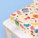 A square booster cushion with a colourful cartoon food and drink print, resting on a white chair against a light blue background.