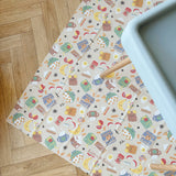 Food-patterned splash mat on a wooden floor with sage green highchair tray partially visible