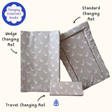 Three changing mats. Wedge, standard and a travel changing mat with a repeating white bird pattern on a warm beige background, labelled with Bobbin and Bumble on a light cream background. A blue circular badge in the top left reads 'Changing Essentials Bundle'.