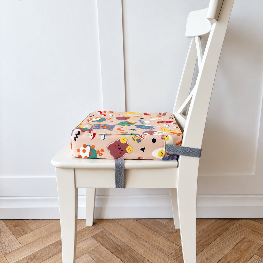 A colorful snack time print booster cushion for toddlers with grey straps, shown on a white wooden chair in a room with a light wood herringbone floor and white paneled wall.