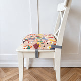 A colorful snack time print booster cushion for toddlers with grey straps, shown on a white wooden chair in a room with a light wood herringbone floor and white paneled wall.