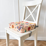 A square booster chair cushion with a colourful cartoon snack print sits on a white wooden chair with a grey strap, set against a white paneled wall and light wood herringbone floor.