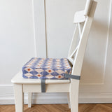 A square fabric booster chair cushion with a blue, white, and yellow diamond pattern, strapped to the seat of a white wooden dining chair on a light wood herringbone floor.