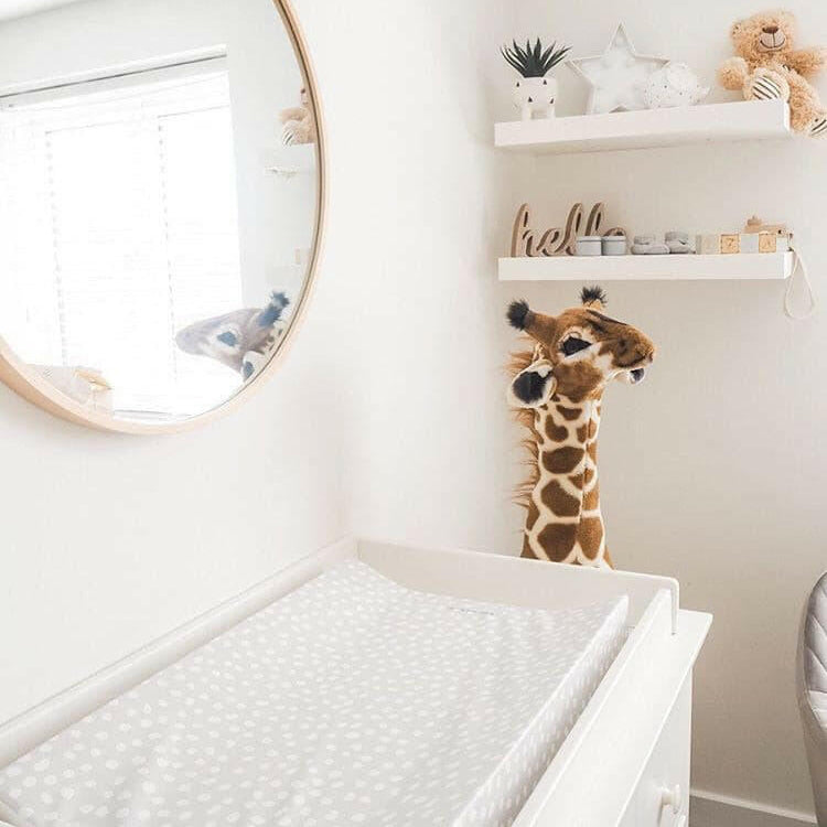 bright nursery with a round mirror on a white wall. nursery shelves with a plush giraffe toy. White changing table with grey spotty changing mat
