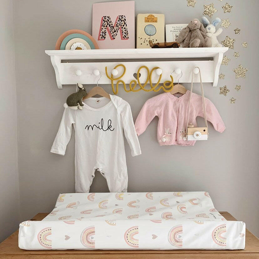 Rainbow print changing mat on top of wooden dresser. Below shelves decorated with baby grows on a grey wall. 