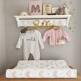 Rainbow print changing mat on top of wooden dresser. Below shelves decorated with baby grows on a grey wall. 